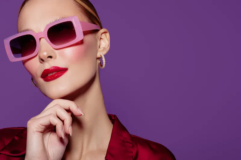 Stylish woman with pink sunglasses and red lipstick poses against purple background. Fashion portrait with glossy makeup and elegant red satin outfit. Concept of glamour and beauty.