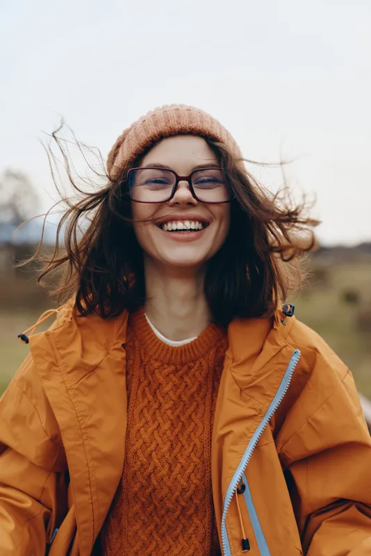 Woman lifestyle outdoors portrait, smiling young woman with glasses and beanie, orange jacket and knit sweater, wind blowing hair, warm autumn light, cheerful casual travel mood