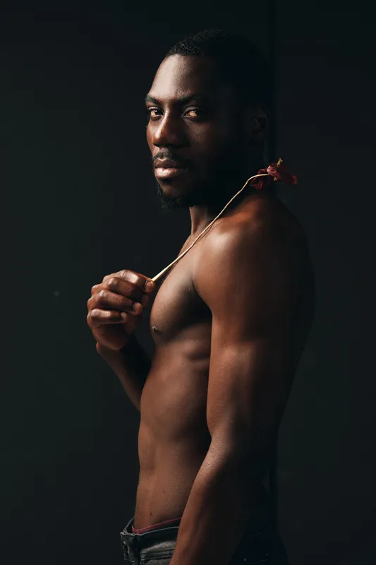 African man with dark skin and muscular body posing shirtless holding a flower on shoulder in creative conceptual visual style with dramatic lighting on black background.