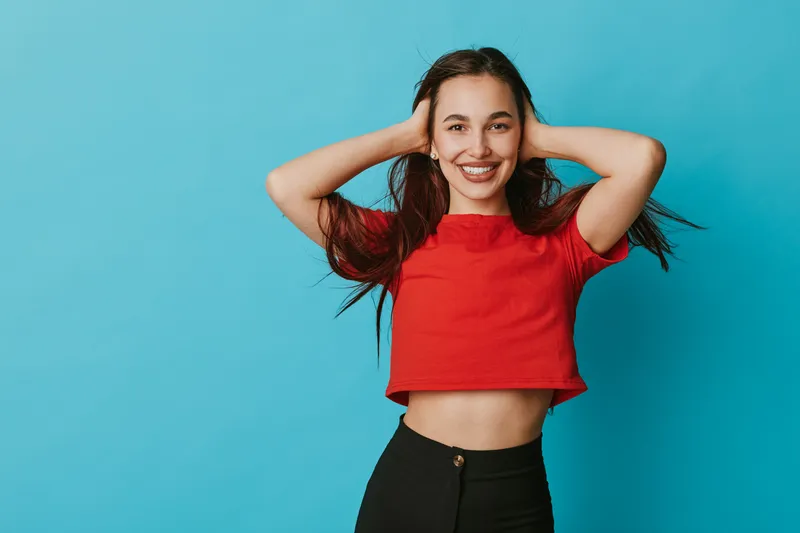 Young woman smiling with hands behind head posing in red crop top and black pants against blue background for casual fashion and happiness concept portrait.