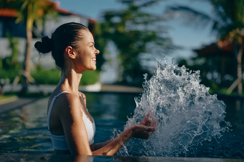 Smiling woman by a pool splashing water, outdoor summer scene, lifestyle moment of wellness and leisure. Bright sunshine and blue tones create a warm, carefree mood.