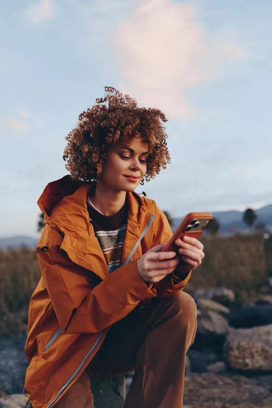 Woman lifestyle smartphone outdoors curly jacket portrait smiling, young female holds a phone while sitting in a countryside field, warm casual travel mood, blue sky and calm nature
