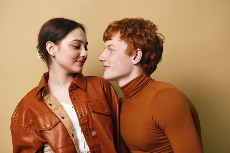 Couple, young man and woman looking at each other with smile and intimacy, wearing brown and orange clothing against plain background. Relationship and affection concept for love and connection.