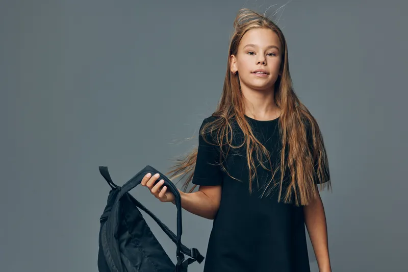Girl with long hair holding black backpack wearing black dress on gray background, standing and looking at camera with neutral expression in studio. Child portrait with casual style and school item.