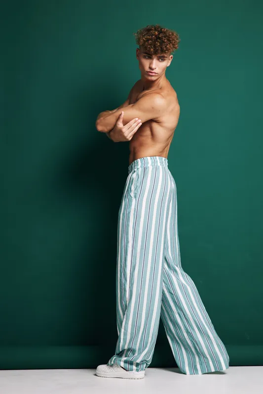 Young man with curly hair posing shirtless in striped wide pants and white sneakers against green background. Fashion model standing with crossed arms and confident expression.