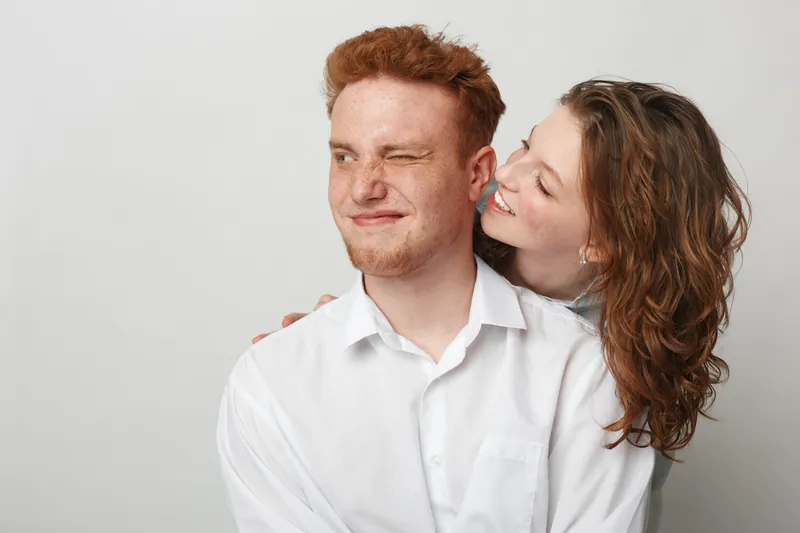Young couple in white shirts share a playful moment, woman smiling and hugging man who winks. Studio portrait on light background, concept of love, friendship, and happiness.