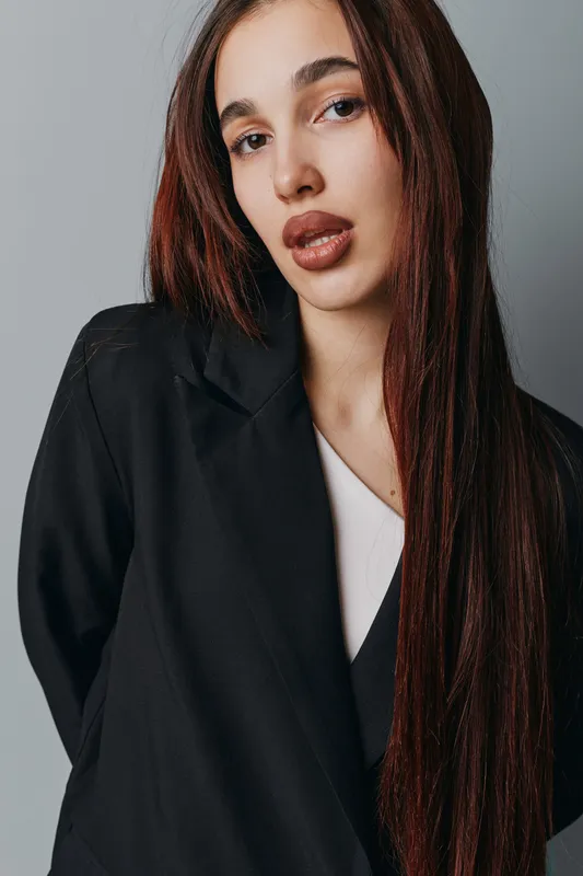 Portrait of confident woman with long hair wearing black blazer and white top standing against gray background. Fashion model with natural makeup and serious expression looking at camera.