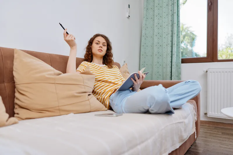 Young woman lounging on a sofa at home, casually holding a remote while resting a notebook on her lap, smiling with a relaxed, focused vibe under warm daylight, striped yellow top, jeans and cushions.