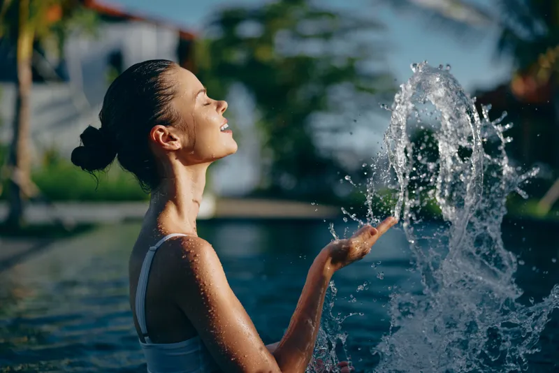 Young woman enjoys sunny day by the pool splashing water with hand, has relaxed positive mood, outdoor lifestyle, natural beauty and summer leisure concept.
