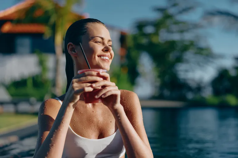 Smiling woman in a white swimsuit enjoying a refreshing beverage beside a sunny pool, capturing outdoor leisure and wellness vibes, with bright lighting that creates a relaxed, cheerful mood and