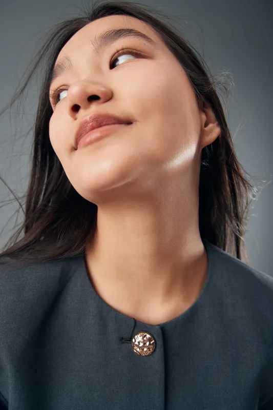 Portrait of young woman looking up with long hair and minimal makeup, wearing a dark top with a decorative button, studio lighting highlighting skin texture and facial features.