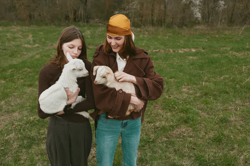 Two women outdoors, smiling as they cradle young goats in their arms, wearing rustic clothing. A candid lifestyle moment highlighting gentle bonding between people and animals in a grassy field.