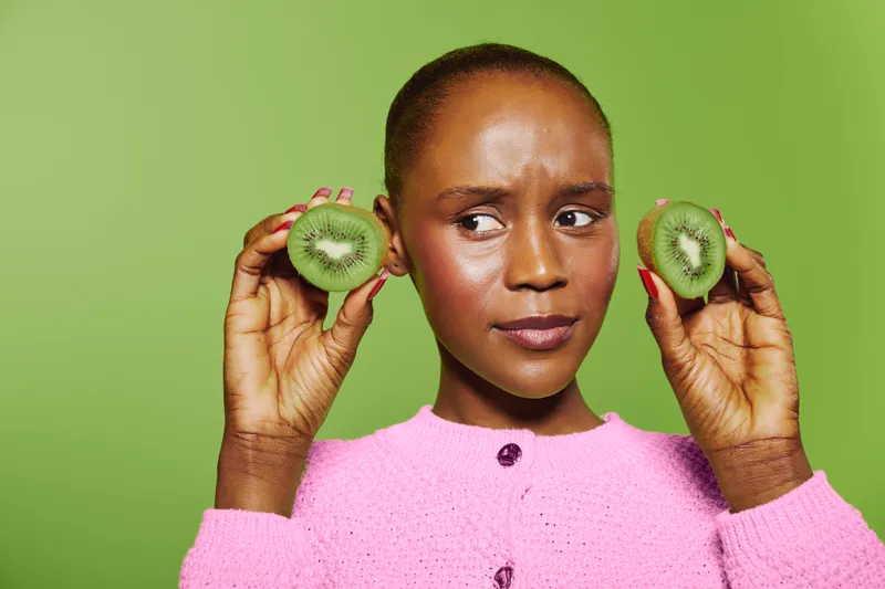 Young African woman with dark skin holds two halves of fresh kiwi near her ears, wearing pink sweater. Green background highlights healthy fruit concept and natural beauty with playful expression.