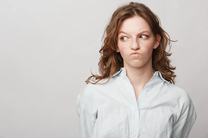 Young woman with curly hair in white shirt looks sideways with skeptical expression on light background. Concept of doubt, suspicion, and uncertain emotions.