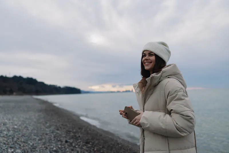 Woman along a pebbled beach in a padded winter coat and knit hat, smiling softly as the sea and cloudy sky frame a serene coastal scene in chilly weather.