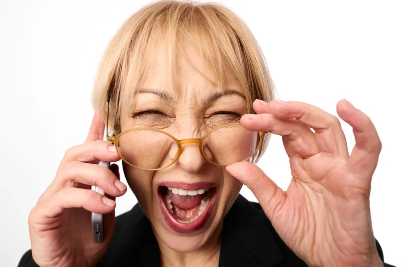 Blonde woman closeup portrait laughing excited, holding glasses with golden frames, eyes behind lenses, one hand near phone, isolated on white background studio
