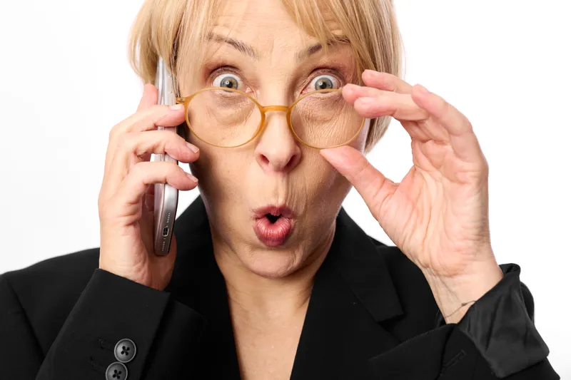 Surprised blonde woman in a business suit holds yellow glasses with one hand and a phone receiver to her ear, mouth open on a white background, shock and stress, instant panic.