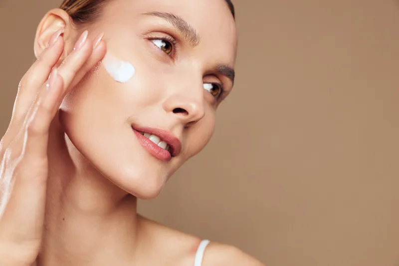 Young woman applies moisturizer to her cheek in a close up studio shot, conveying a calm skincare routine and confident, serene mood.