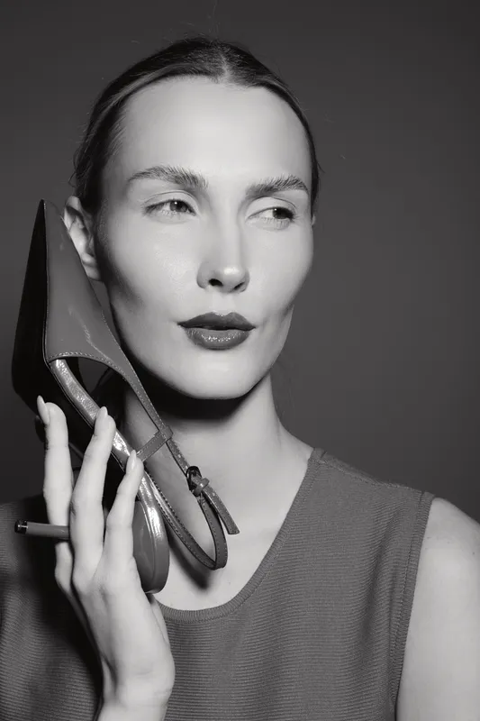 A pale skinned caucasian woman with slicked back hair holds a high heel shoe near her face, posing confidently in a minimalist studio setting. Concept of fashion and elegance.