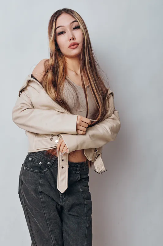Young woman posing with beige jacket off shoulder and casual outfit standing against light gray background with confident expression for fashion portrait.