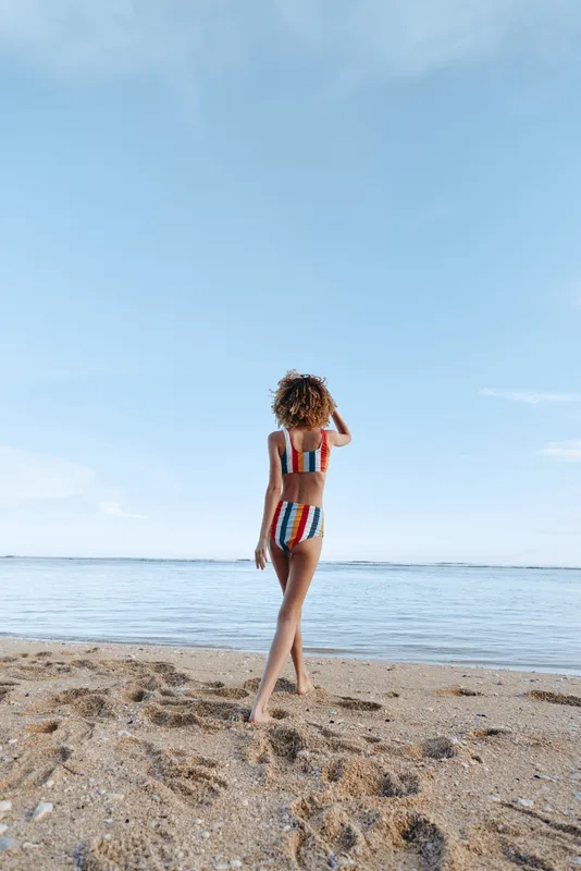 Beach scene shows a woman in a striped bikini walking along the sand with the calm ocean in the background, capturing a relaxed, sunny summer mood, soft breeze, and casual beachwear style.