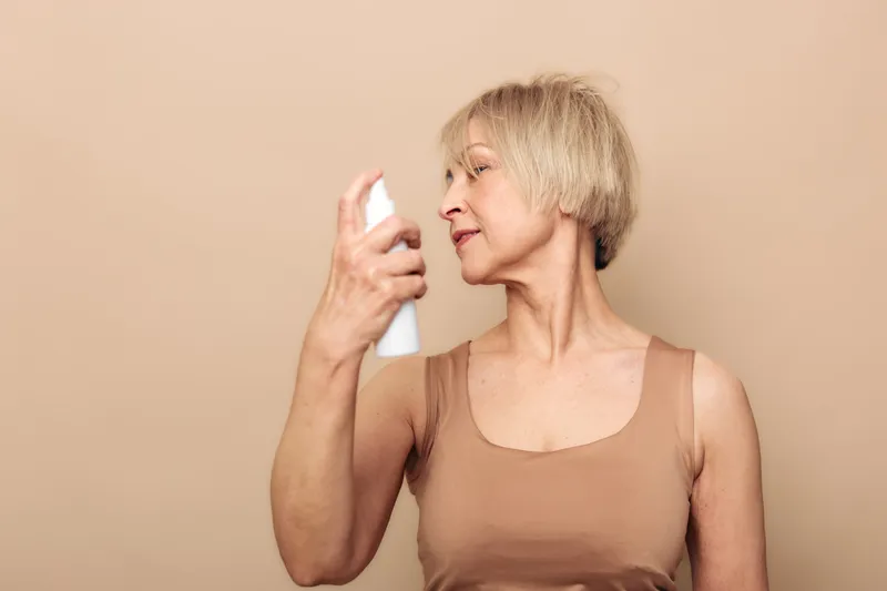 Mature woman with short blonde hair sprays mist on her face wearing beige tank top. Neutral background with soft lighting highlights skincare routine and self-care for aging skin.