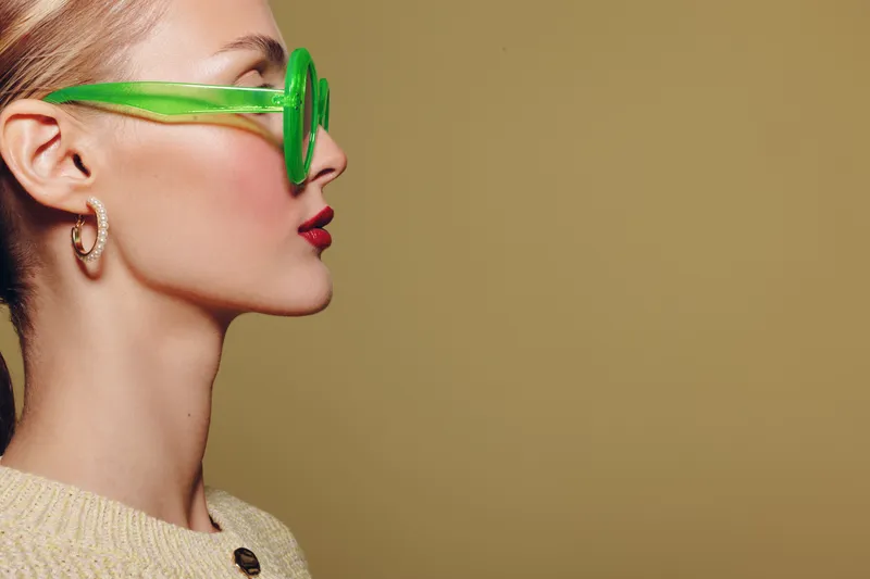 Profile of a young woman wearing bright green sunglasses and a beige sweater against a plain background. Concept of fashion, style, and modern accessories.