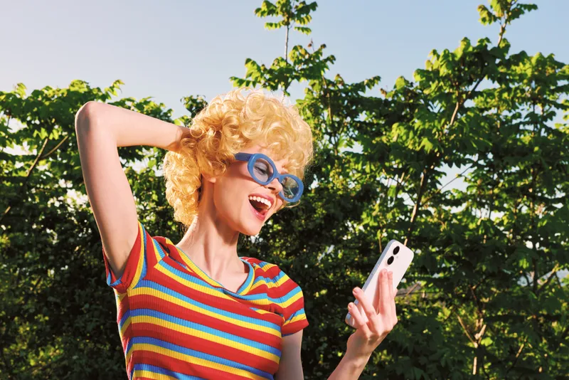 Cheerful woman outdoors in a striped shirt, laughing with sunglasses, capturing a sunny moment, relaxed pose, retro film color palette, lush greens in the background