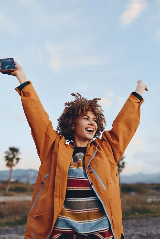 Woman lifestyle outdoors, orange jacket, curly hair, smiling, arms raised while holding a smartphone under open sky. Relaxed travel moment in nature, warm light, distant mountains, casual portrait.