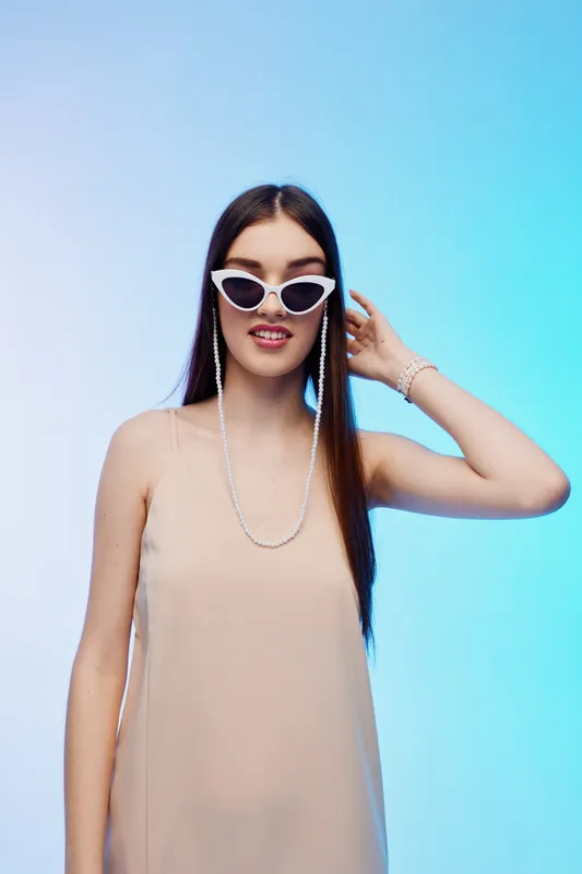 Young woman in beige dress wearing white cat eye sunglasses and jewelry poses with hand near face on blue gradient background. Concept of woman beauty fashion style.