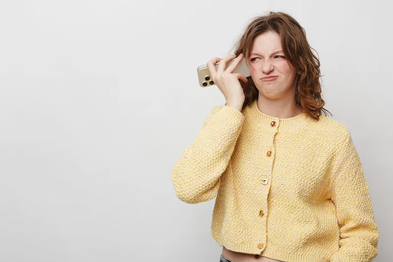 Young woman in yellow sweater holds smartphone to ear with annoyed expression on white background. Concept of frustration, bad phone call, and communication problems.