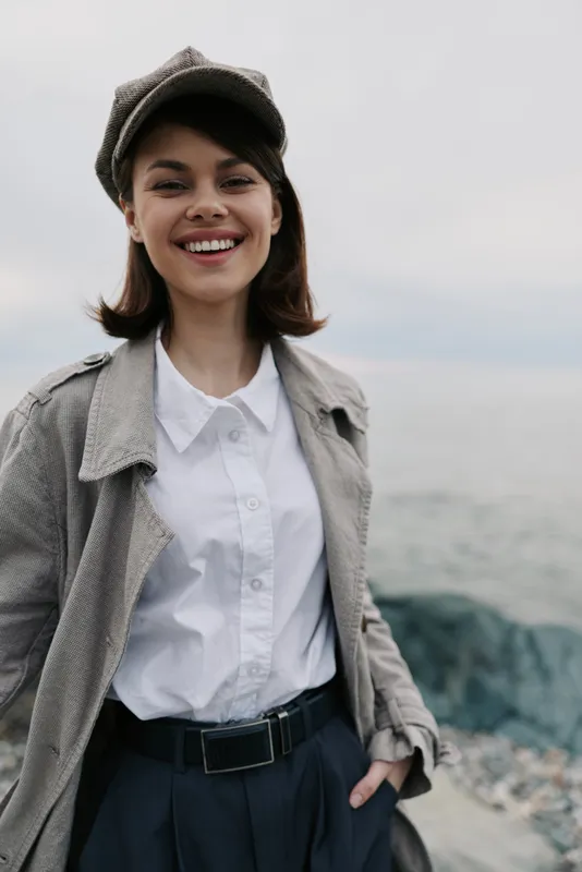 Smiling young woman in casual stylish outfit stands near rocky coast. She wears a hat, white shirt, and light jacket with relaxed posture, enjoying outdoor fresh air and nature.