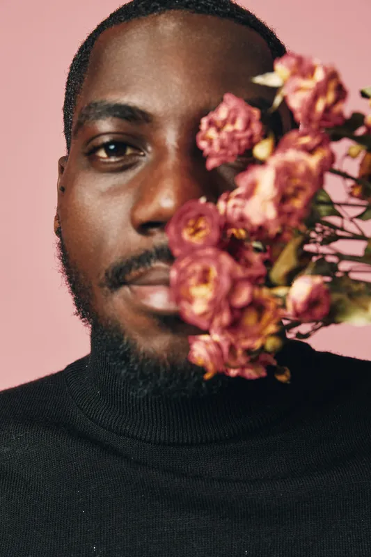African man with dark skin and beard wearing black sweater holding dried pink flowers close to face on pink background in creative conceptual visual style portrait.