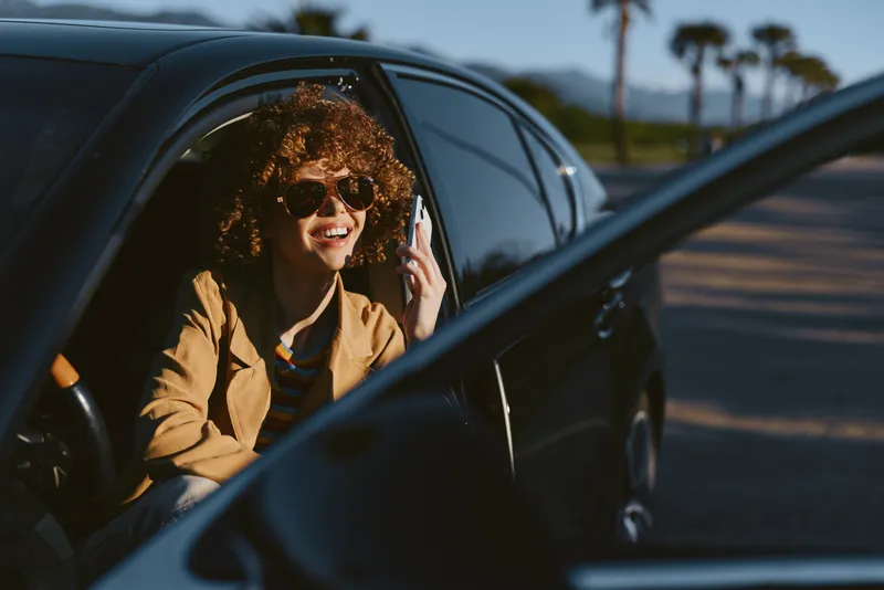 Woman with curly hair wears a brown blazer, sits in an open car window, smiles behind sunglasses, enjoying a casual street moment with a sunny outdoor backdrop and retro film color vibe.