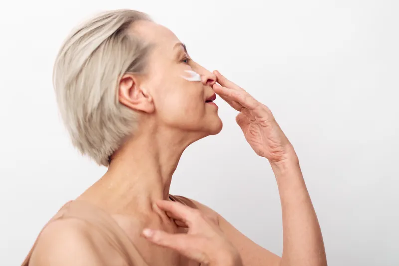 Mature woman with short grey hair applies face cream on cheek in bright studio. Concept of skincare, aging skin care, hydration, and natural beauty for elderly women.