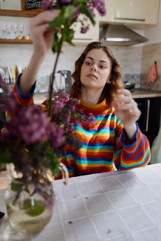 Young woman in a rainbow striped sweater arranges a bouquet at a bright kitchen table, a calm domestic moment captured with natural light and soft colors.