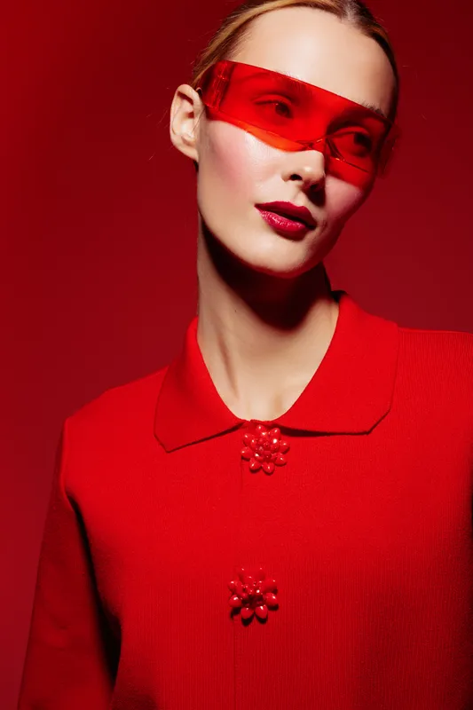 Portrait of a stylish woman wearing red sunglasses and a red textured jacket against a red background. Fashion concept with bold makeup and modern accessories.