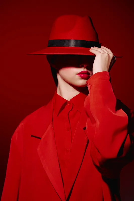 Fashionable woman in bold red coat and hat partially covers her face, wearing dark lipstick. Creative beauty style against a rich red background emphasizes glamour and mystery.