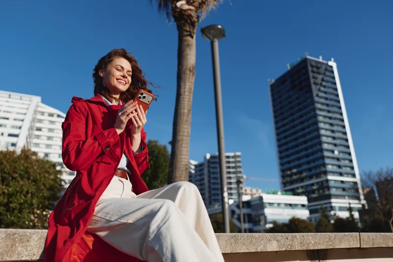 Young woman in a red coat smiles while using a smartphone outdoors on a sunny day with urban buildings in the background.