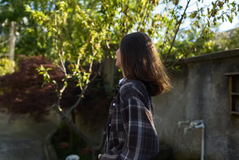 Candid woman in plaid shirt walks through a sunlit garden, capturing a simple lifestyle moment with calm, reflective mood and natural surroundings