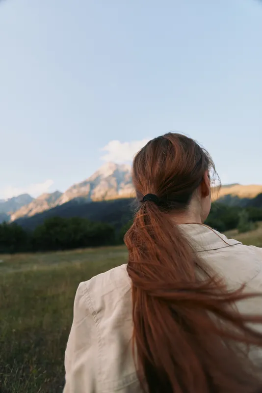 Young woman with long hair tied in ponytail stands outdoors looking at mountain landscape during clear day, enjoying peaceful nature and lifestyle moment.