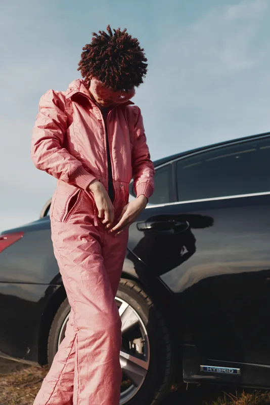 Pink jumpsuit, car, outdoor fashion and street style portrait of a person adjusting sleeves beside a sleek vehicle, urban vibe, confident pose, lifestyle photography with daylight and modern attitude.
