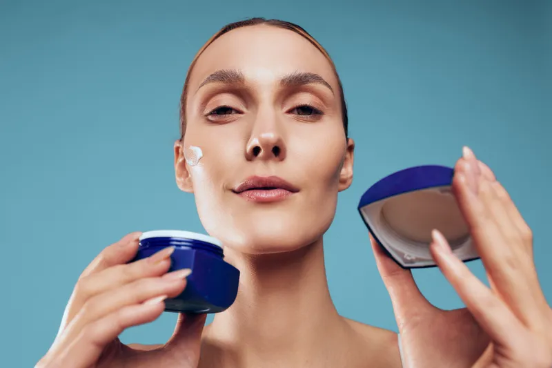 Young woman with brunette hair applies moisturizing cream on her cheek while holding a blue jar against a plain blue background in a skincare routine.