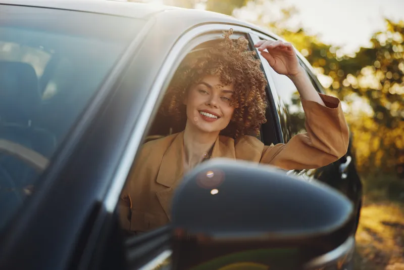 A smiling woman leans out of a car window in warm sunlight, capturing a casual lifestyle moment. Vintage color tones emphasize carefree travel and everyday joy.