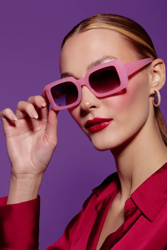Stylish woman with pink sunglasses and red lipstick poses against purple background. Fashion portrait with trendy accessories and vibrant colors for beauty and style concept.