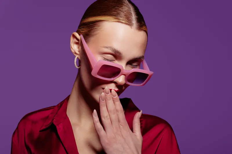 Young woman with slicked hair wearing pink sunglasses and red shirt winks and covers mouth with hand on purple background. Concept of fashion, style, and playful attitude in studio.