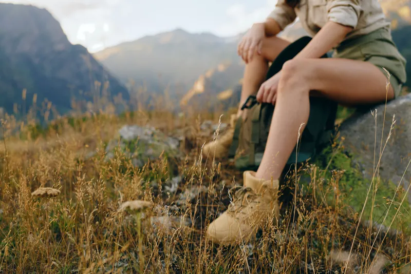Woman wearing hiking boots sitting on a rock in a mountain meadow. Outdoor lifestyle concept with nature, adventure, and relaxation during a sunny day.