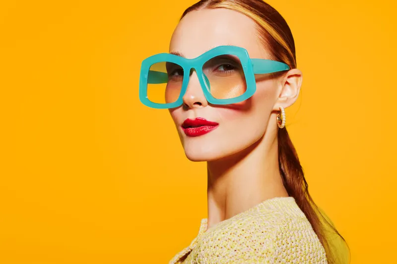Stylish woman with blue oversized glasses and red lipstick poses against bright yellow background. Fashion portrait with modern accessories and confident expression.