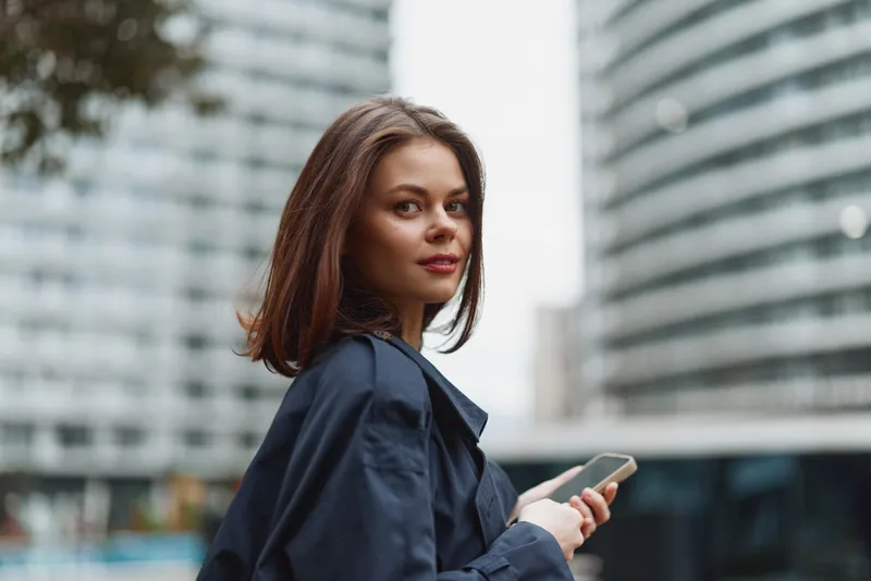 Young woman with brown hair holds a smartphone while standing outdoors near modern glass buildings, looking confidently at the camera with a slight smile.