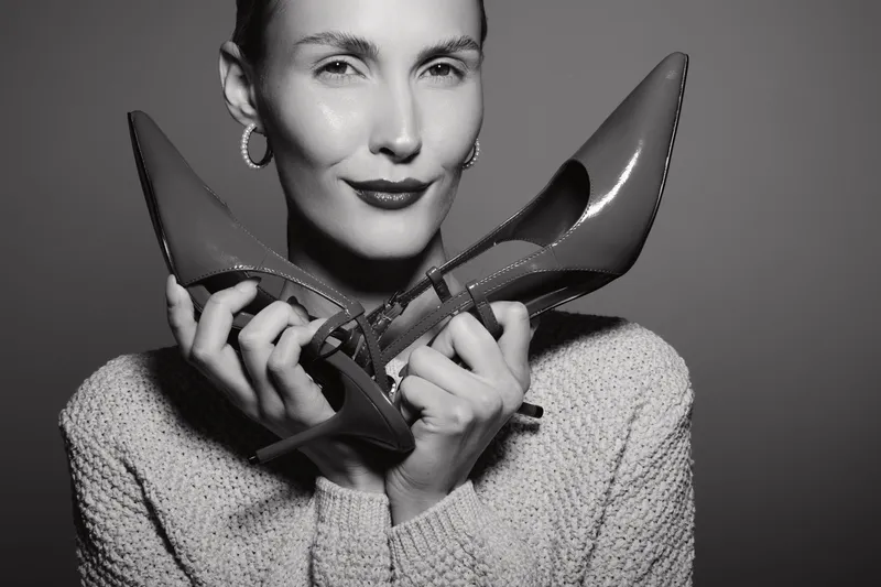 Young white woman with light skin holds pair of high heel shoes near face with gentle smile. Studio portrait in black and white. Concept for fashion, style, and elegance.